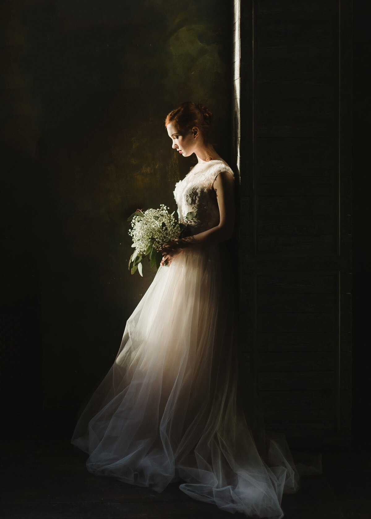 A red headed bride leans against a wall in her wedding dress, holding a beautiful bouquet.