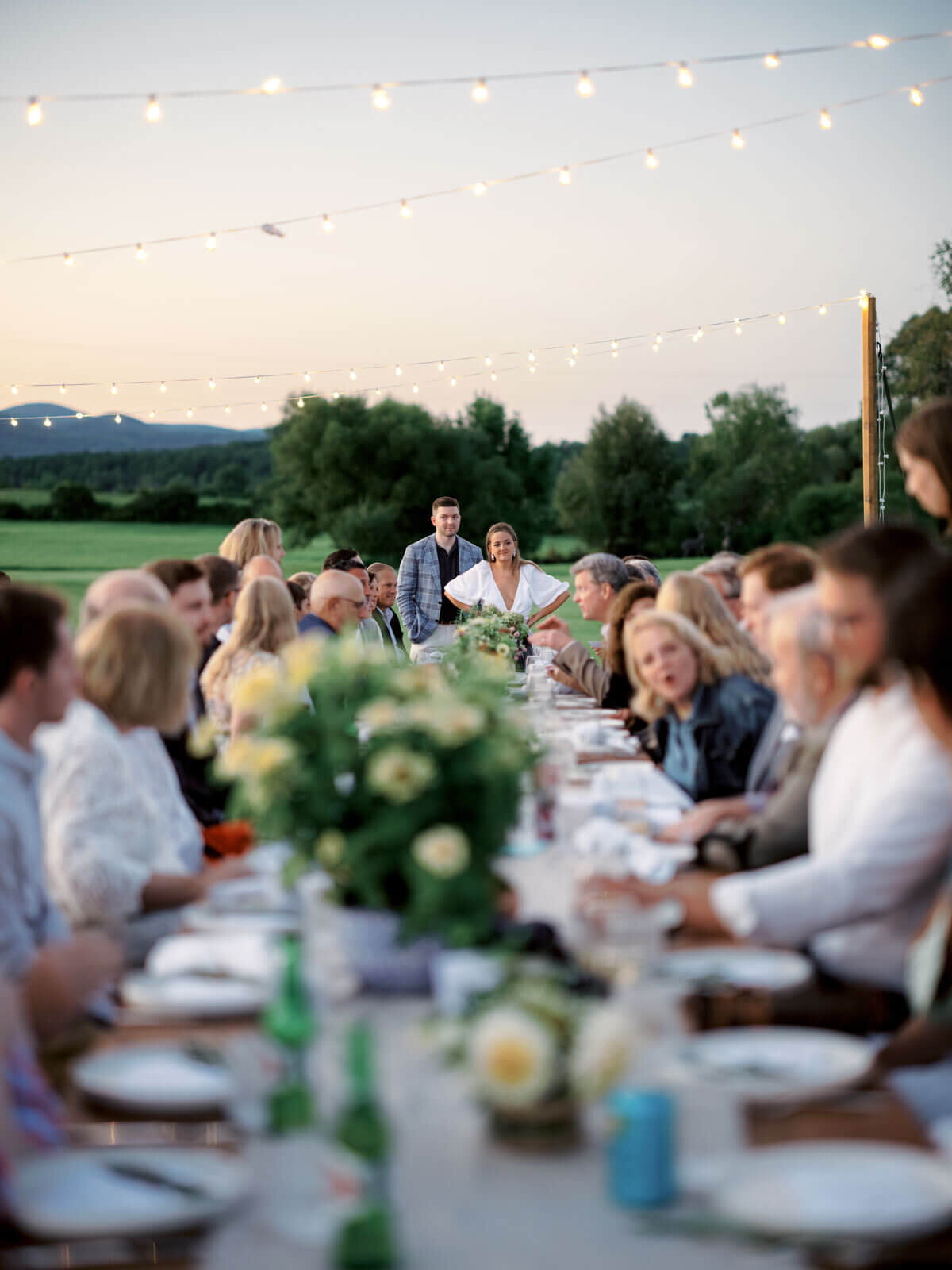 The bride and groom-to-be are with guests seated on a long dining table outdoors at Lion Rock Farms, CT. Image by Jenny Fu Studio