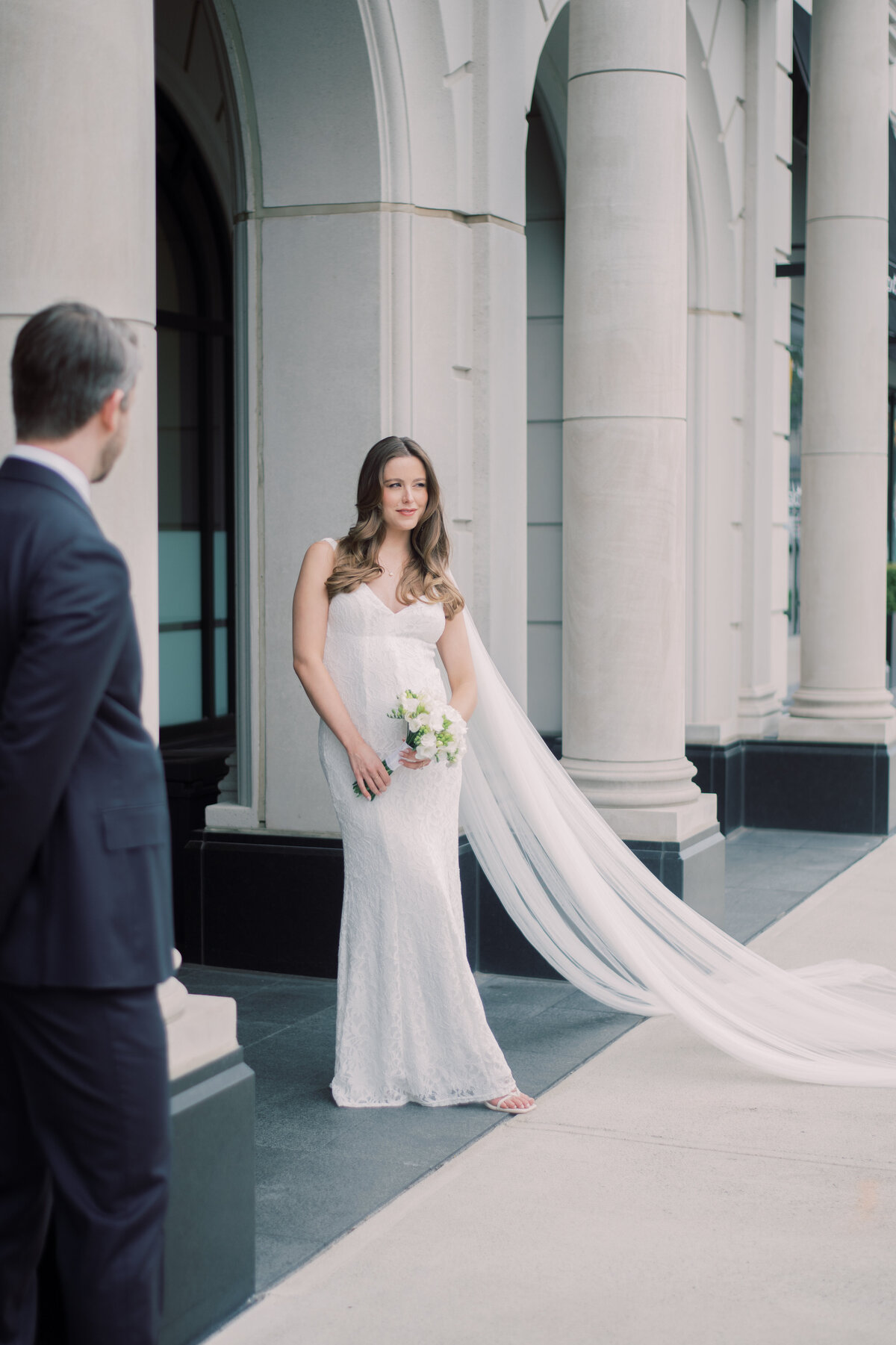 Bride poses on the pillar while groom watches her.