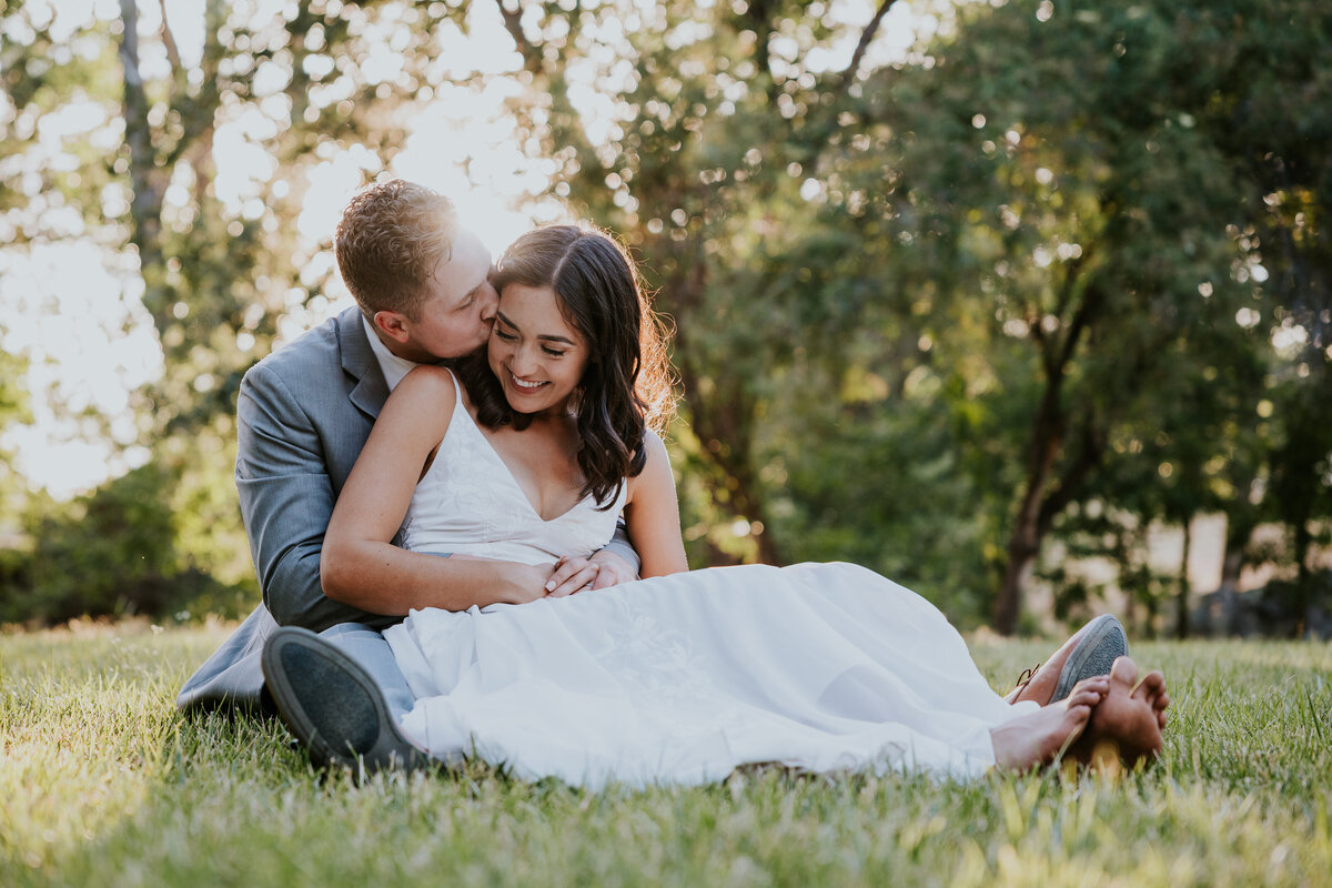 Bride and groom snuggle together on grass while groom kisses brides cheek.