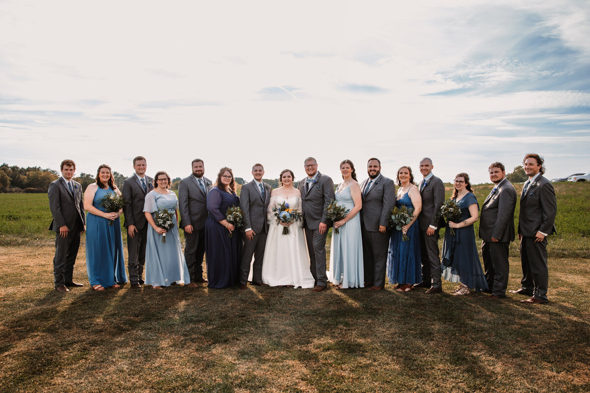 A large wedding party stands in a field with the newlyweds in the middle