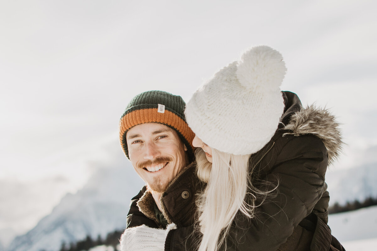 man smiling at the camera as his fiance looks at his for their winter engagement session in Jackson Hole Wyoming