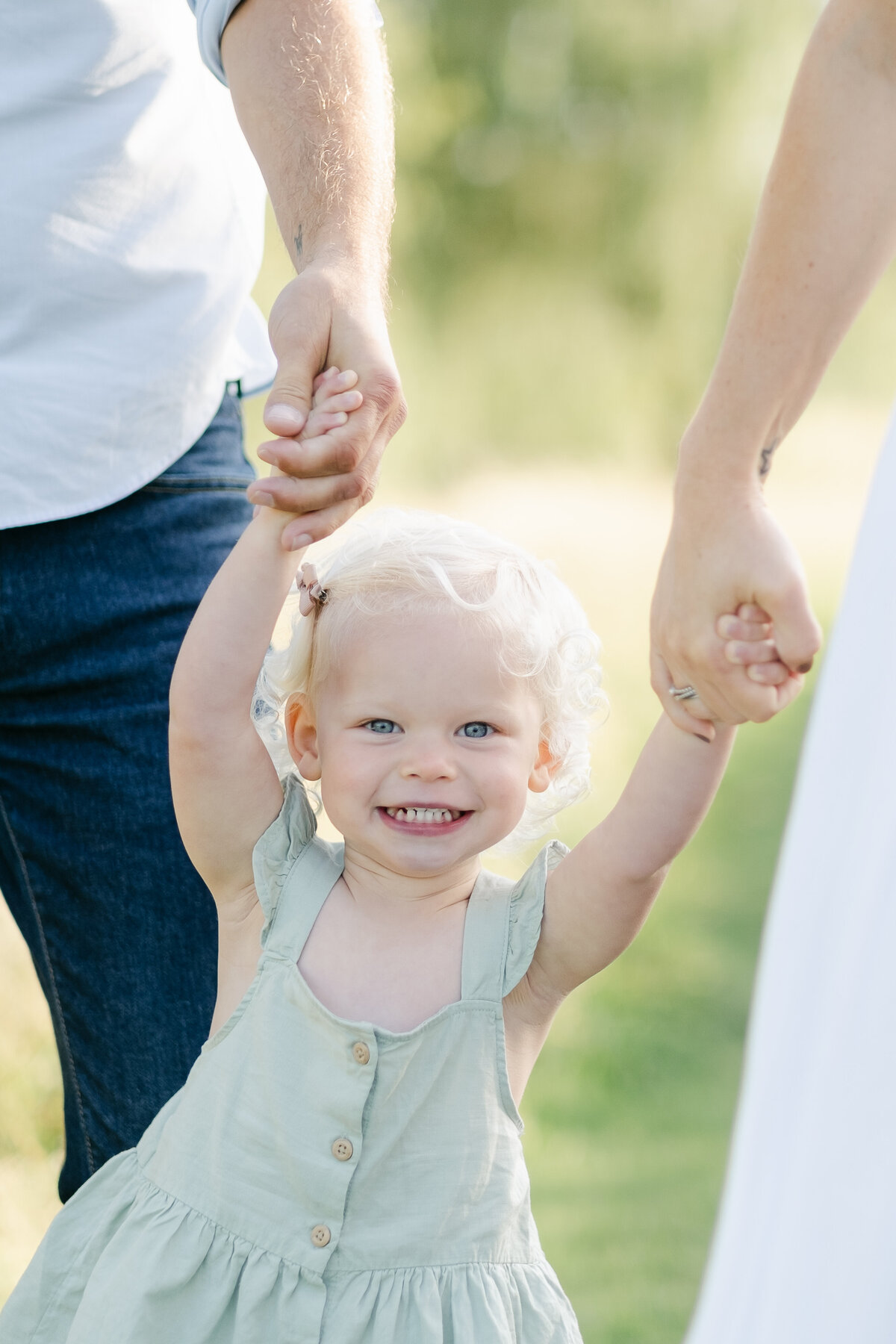 Little girl smiles at the camera while her parents hold her hands