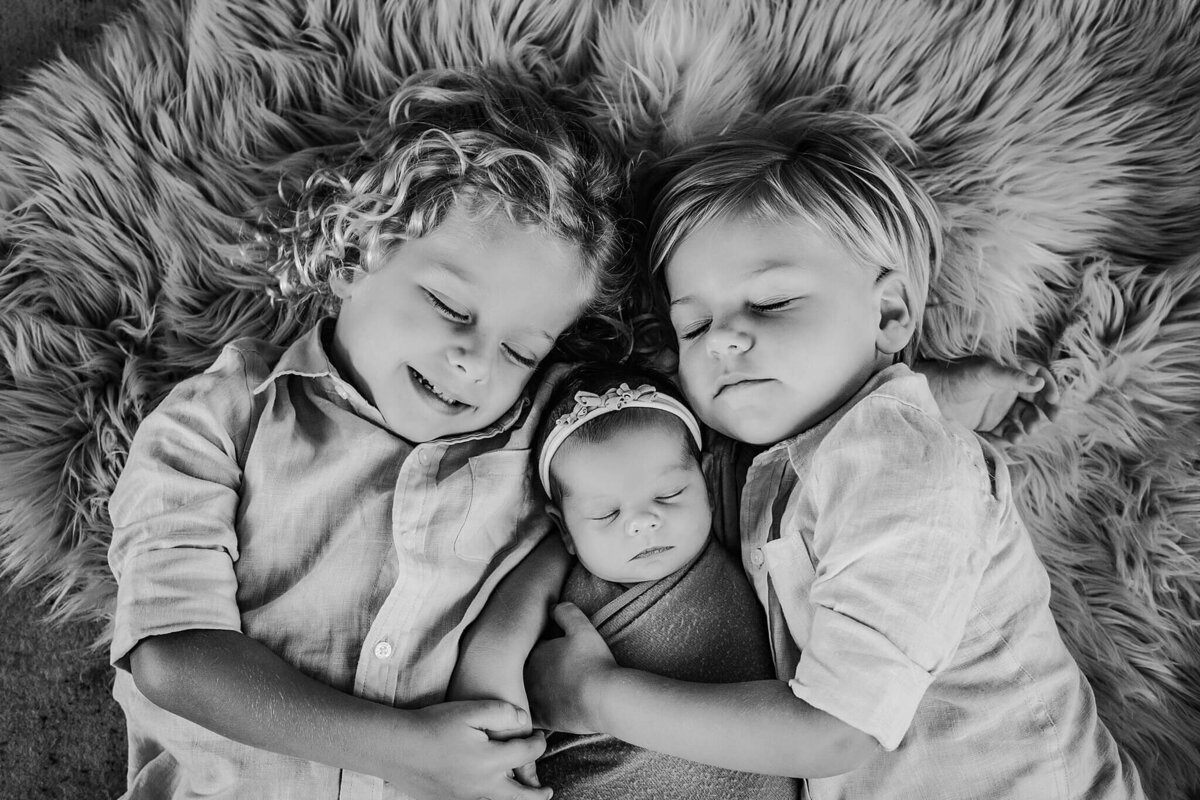 A black and white photo of three siblings lying on a furry rug. The two older siblings, a boy and a girl, are lying on either side of the baby, both with their arms wrapped around her. The older siblings are both looking at the camera, while the baby is sleeping peacefully. The photo is a heartwarming depiction of sibling love and protection. The soft focus and muted tones contribute to the overall sense of calm and tranquility. The simple composition and intimate setting make for a touching and timeless image. 