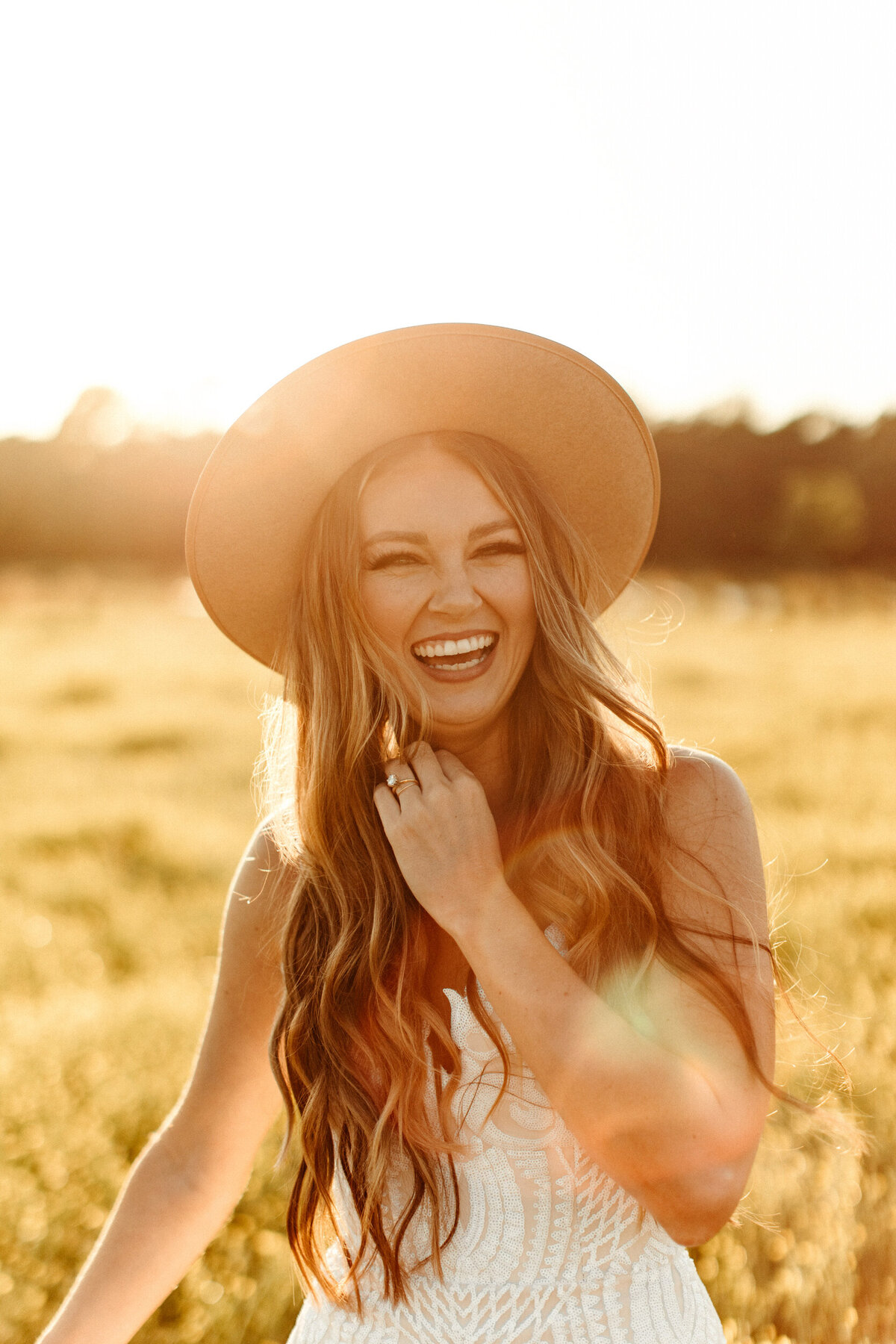 Boho bride in hat running her fingers through her hair and laughing in a field with yellow flowers at sunset
