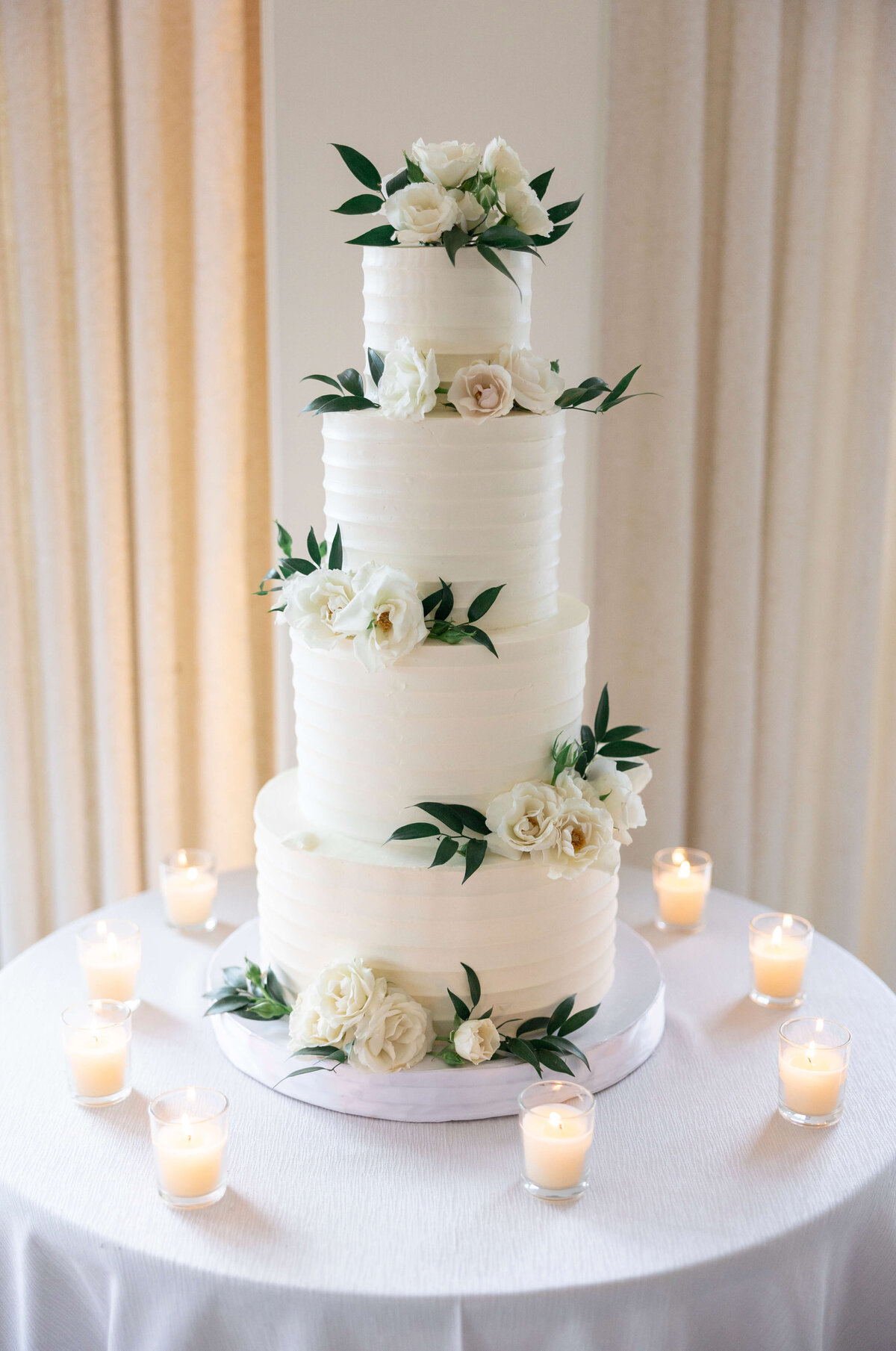 A four-tiered wedding cake decorated with white flowers and green leaves, placed on a white tablecloth. The cake is surrounded by small lit candles, creating an elegant and romantic setting.