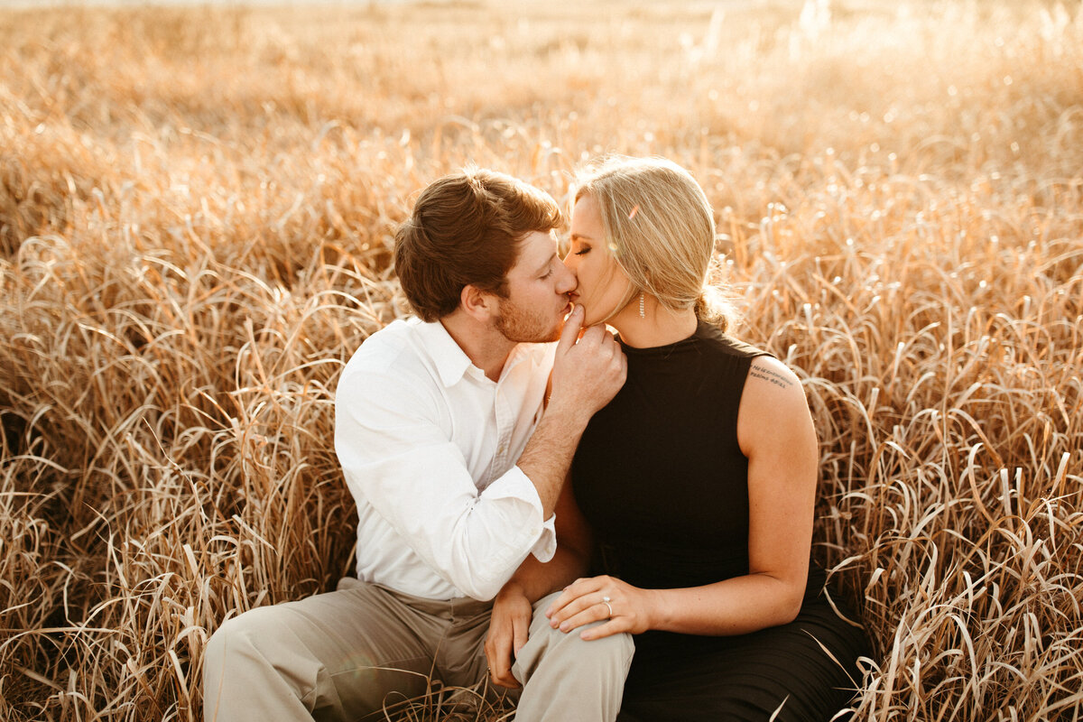 A girl in a fitted black dress with a messy updo is sitting in a field of tan grass beside her fiancé while he is pulling her in by her chin and kissing her.