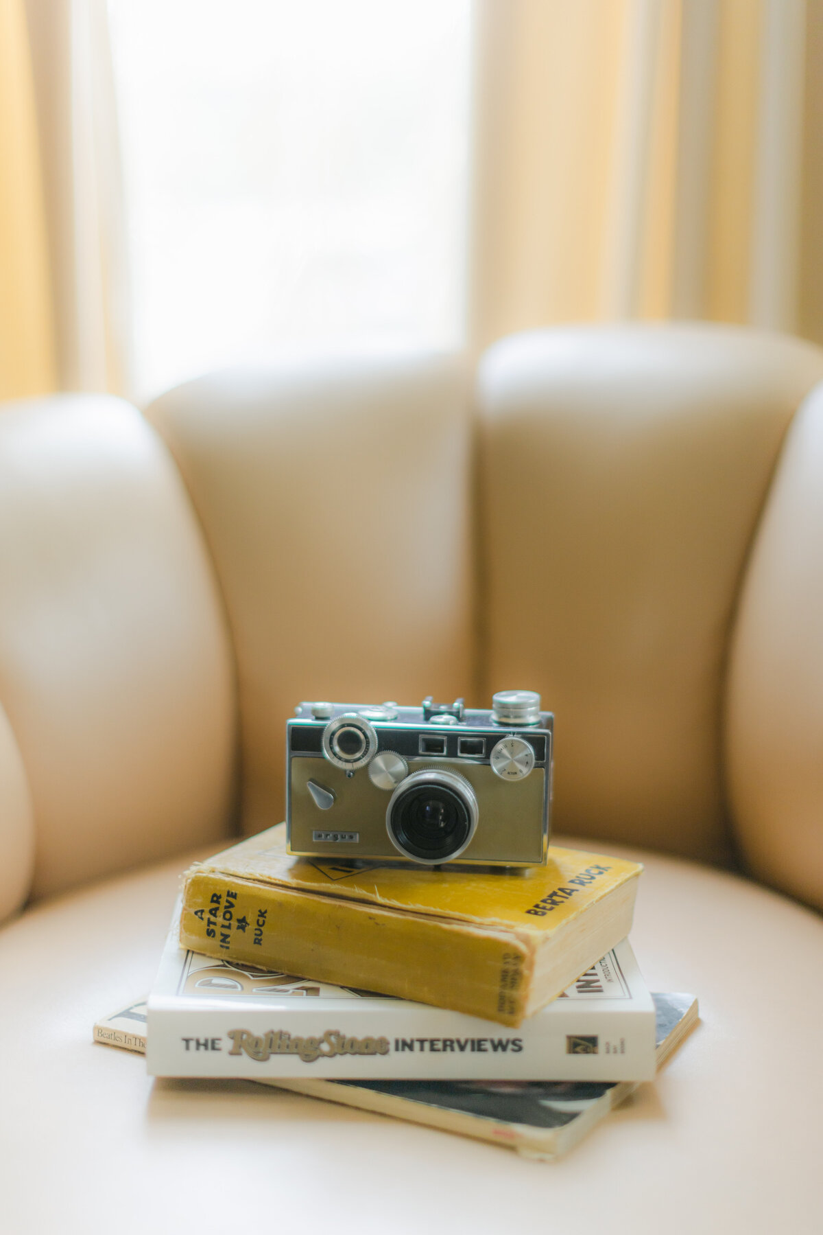 A styled flat lay featuring a vintage Argus camera placed on top of stacked books, including The Rolling Stone Interviews and A Star in Love, all set against a soft neutral armchair and dreamy window light. Captured by Dani Lacey Photographs.