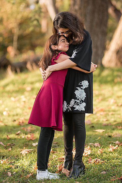 A family of four, dressed in fall colors, together in vibrant foliage of Lincoln Park in West Seattle. The sun filters through the trees at golden hour.  Casting a warm glow on the family. . This photo is a perfect example of fall family mini sessions with Heleyna Holmes Photography,