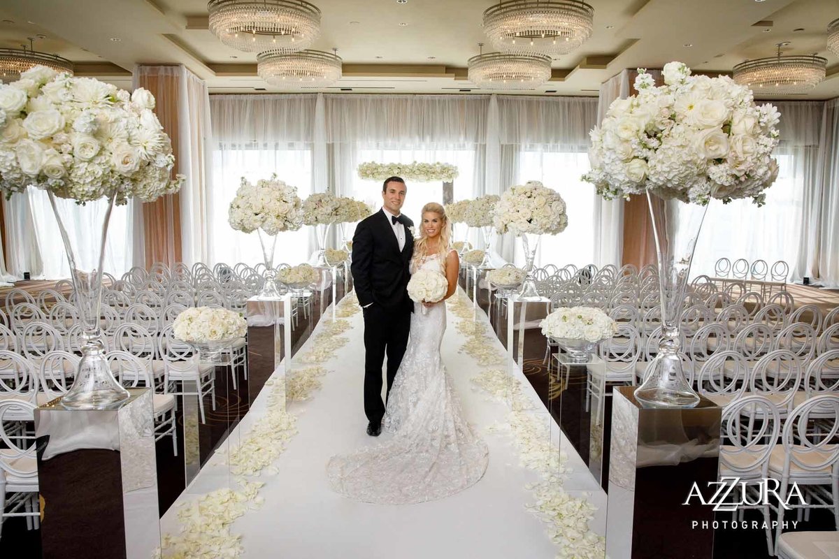 Bride and groom standing in aisle lined with large white flower arrangements, at super Glamorous white Four Seasons Seattle ballroom wedding