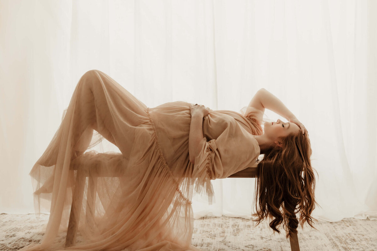 Studio maternity portrait of a mother laying on a bench wearing a peach colored dress.
