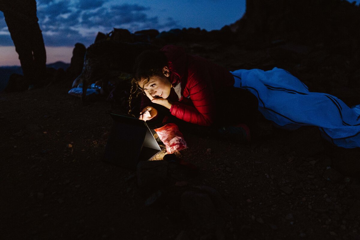 Bride in a red jacket gets ready in a mirror by lantern light before sunrise at the base of Mount Sneffels in Colorado, with her wedding dress draped beside her.