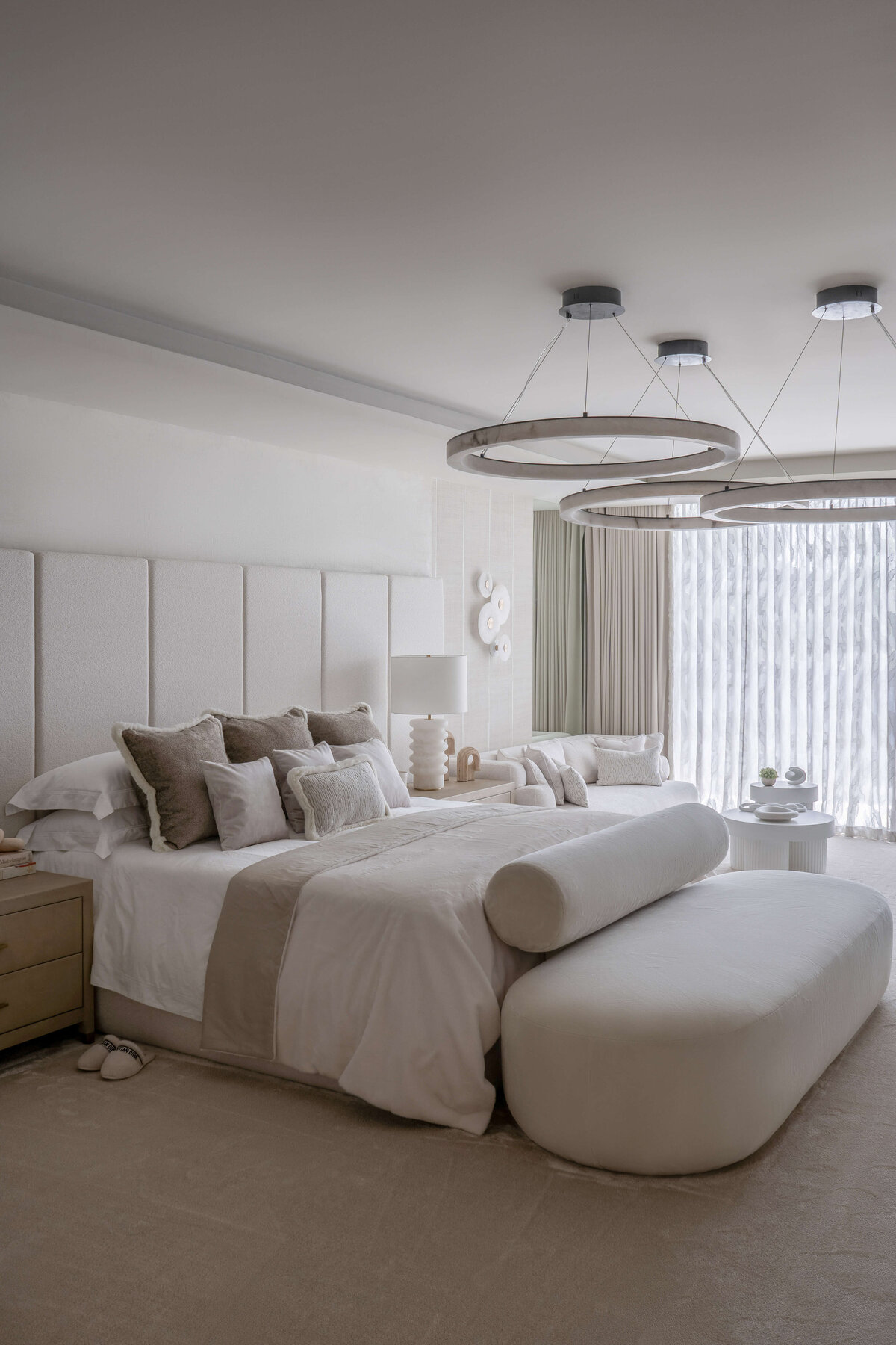 A spacious primary bathroom with a  bright, airy feel. The large, white bed is decorated with neutral accent pillows, with light wood side tables at each side. There is a large white footboard bench, and above the bed is a large, contemporary light fixture. composed of three circles.