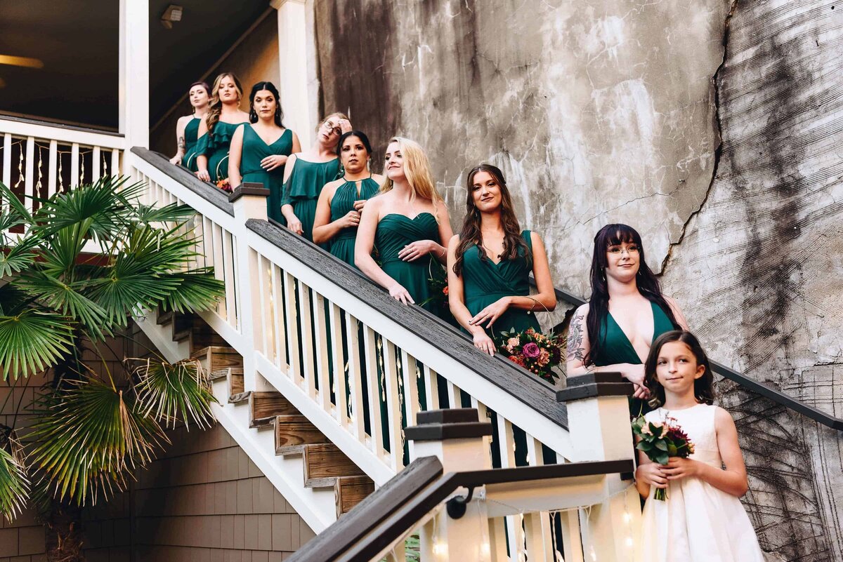 A group of bridesmaids in green dresses and a young flower girl in white stand on a staircase in an outdoor venue in Lincoln Parish. Beside them is a weathered wall. They are holding bouquets and smiling, with greenery and string lights nearby.