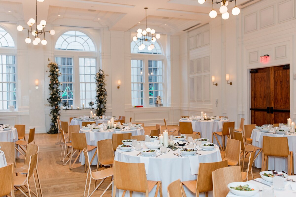 The ballroom at Noelle Nashville is light and bright with ivory walls, tall arched windows and blonde hardwood floors. The room is set with tables covered in white linen set with candles and greenery surrounded by blonde wood chairs. An asymmetrical arbor of greenery with blue hydrangea, white roses and pink roses frames the window.