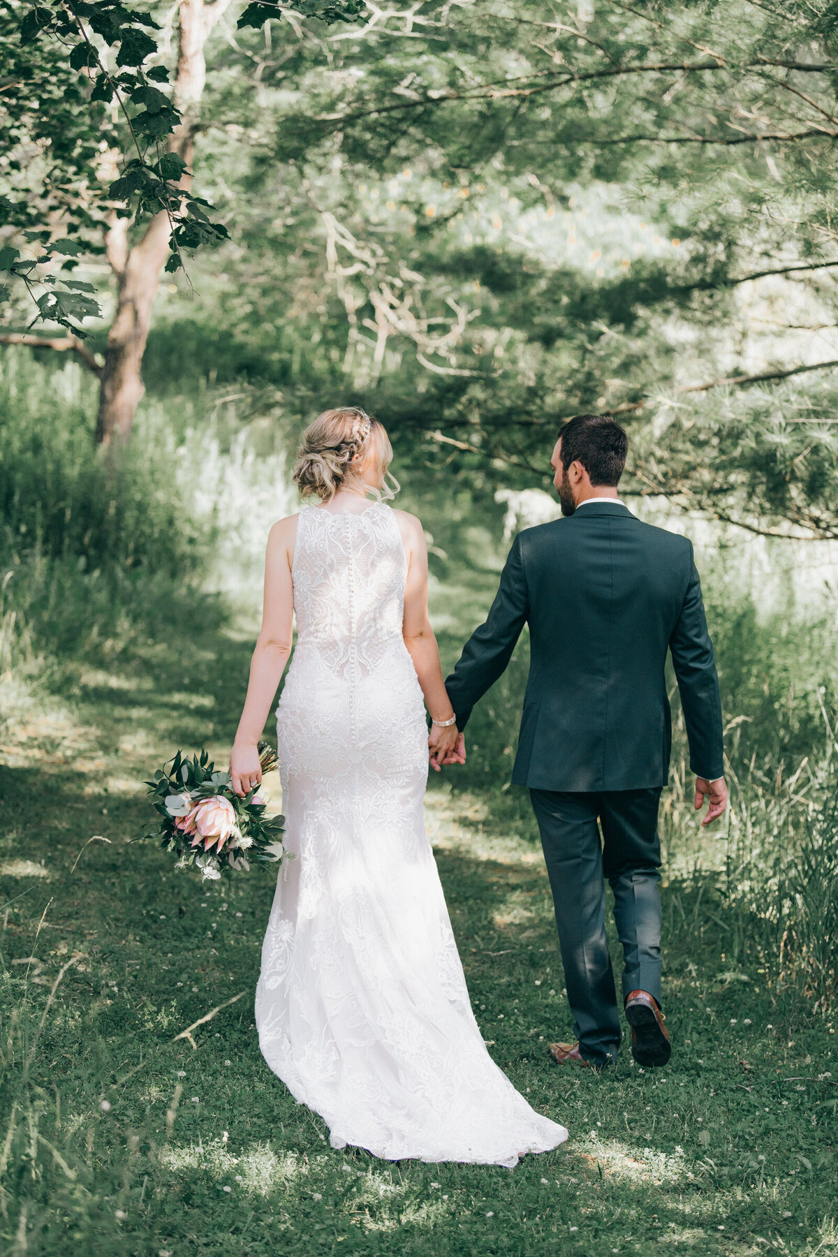 A bride wearing an elegant ivory wedding dress with a beautiful lace back walking hand in hand with her groom wearing a casual green suit