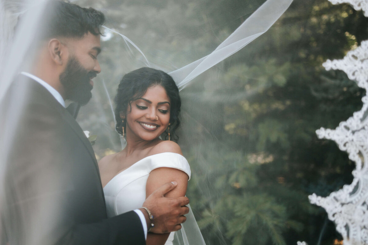 Bride and groom during portraits under veil. Bride is smiling looking to the side while groom is looking at his bride to be
