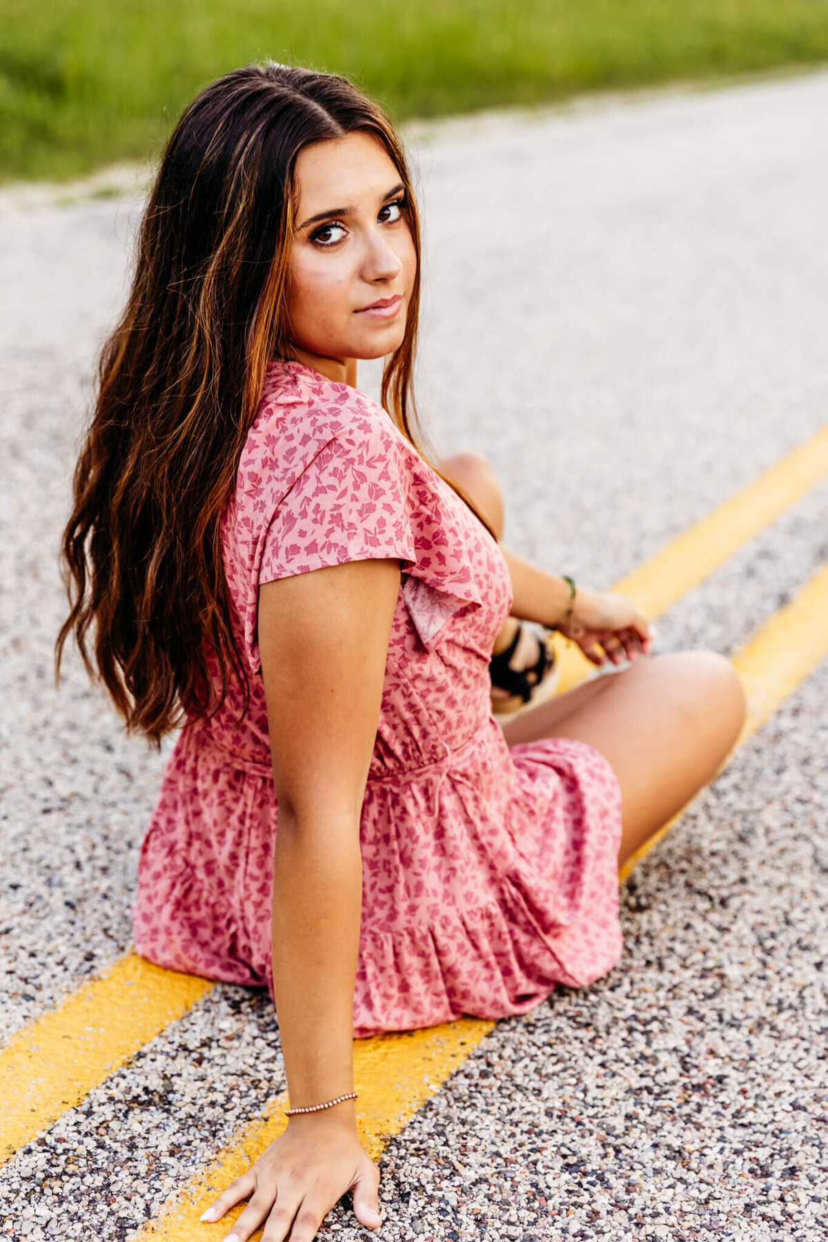 gorgeous high school girl in a pink and red romper sitting on the yellow lines on a road and looking back seriously as she enjoys her Oshkosh senior photography experience.