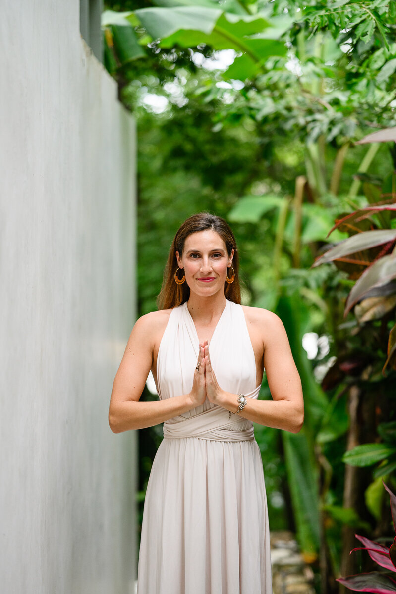A yoga teacher wearing a white dress holds prayer pose while looking at a brand photographer in Denver for her brand photos and brand photography session.
