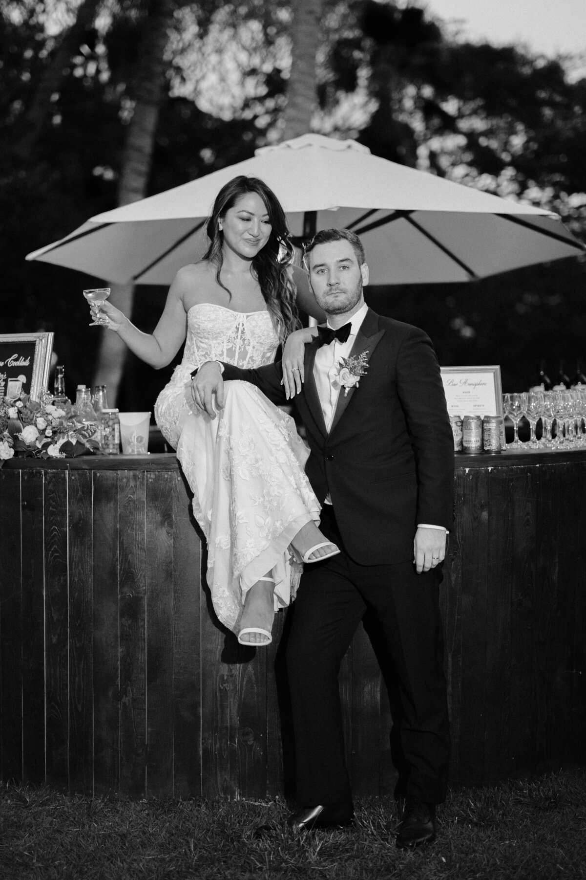 Bride sitting on the dark wood bar looking at groom as he looks to the camera in black and white