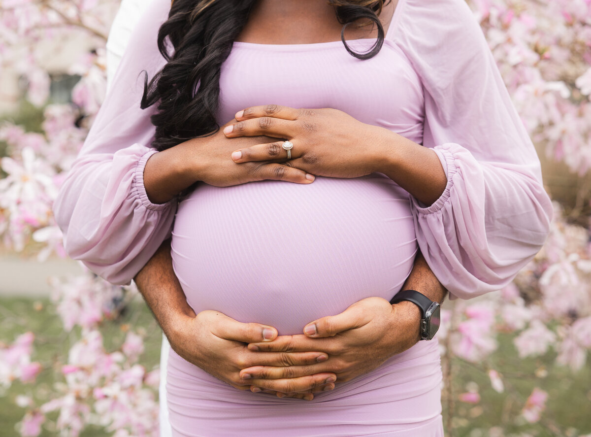 Glowing pregnant woman in a light purple dress gently holding her baby bump and smiling, standing beneath cherry blossom trees during an outdoor maternity photoshoot.