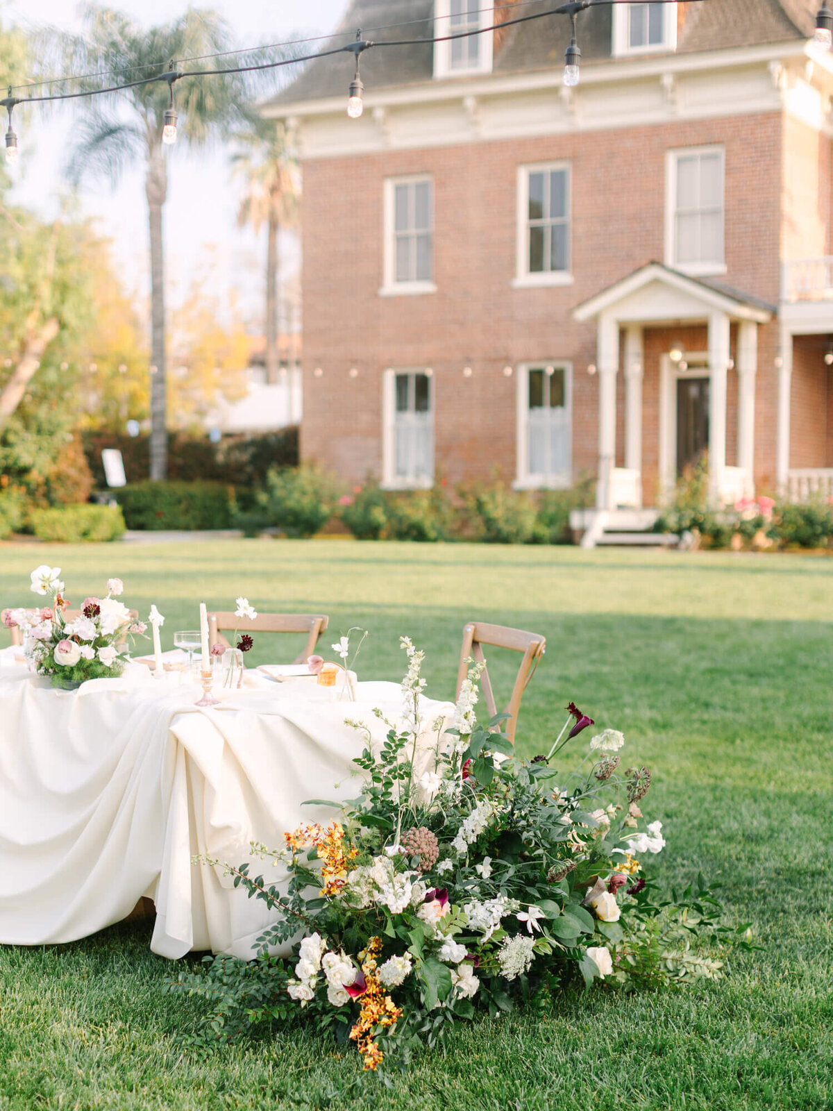 Elegant outdoor wedding setup with a white-clothed table adorned with pink and white flowers, lush greenery, in front of a large brick house. 