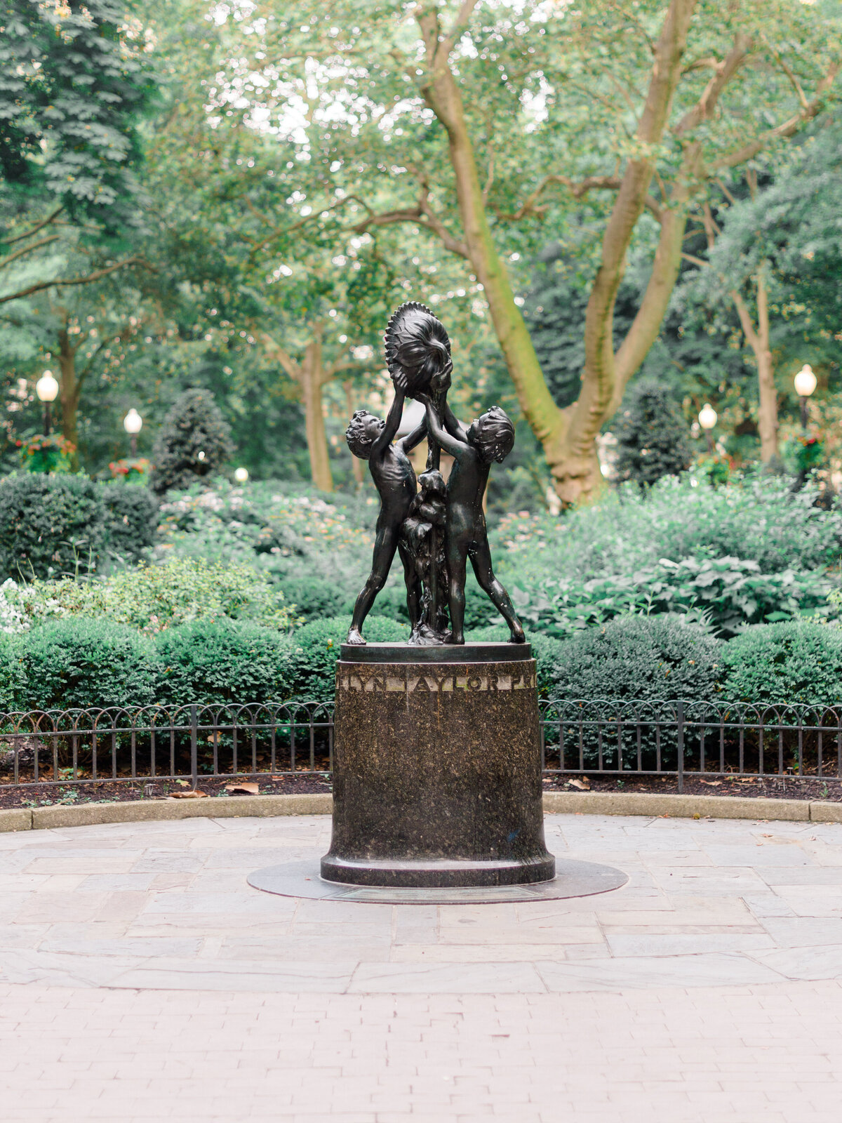 A bronze sculpture of children holding a decorative basin stands at the center of a quiet path in Rittenhouse Square, surrounded by lush greenery and historic charm. Photographed by Elizabeth Kane Photography, a Luxury Philadelphia Wedding Photographer, this image captures the timeless character of one of the city's most beloved parks.