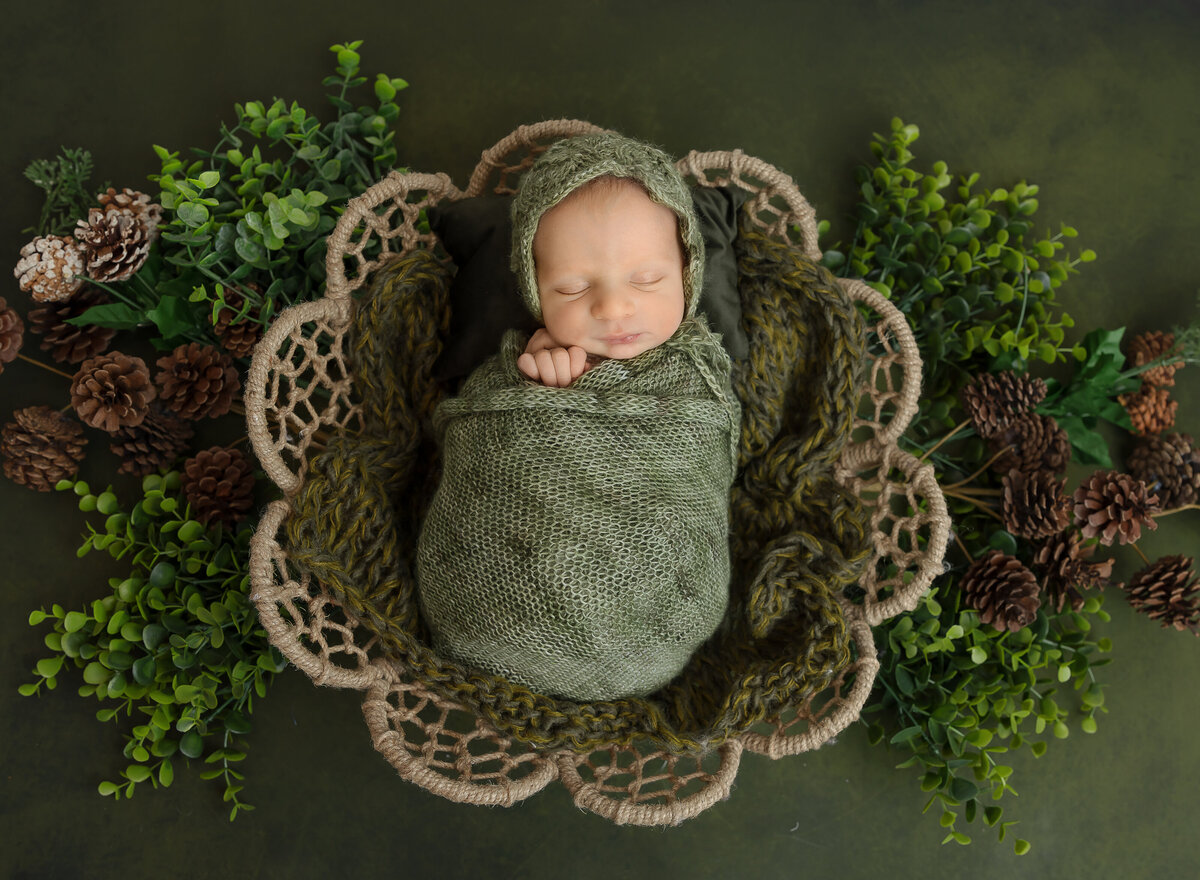 Baby girl swaddled in green is sleeping in a basket for their newborn photoshoot. The basket is placed atop of brown and taupe flowers. Captured by top Brooklyn newborn photographer Chaya Bornstein