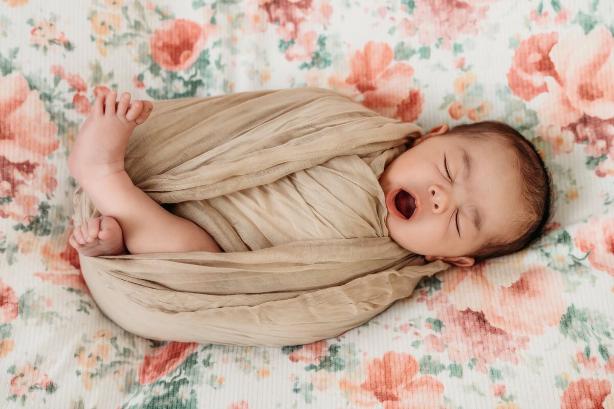 baby in tan swaddle yawning. she is laying on a floral backdrop