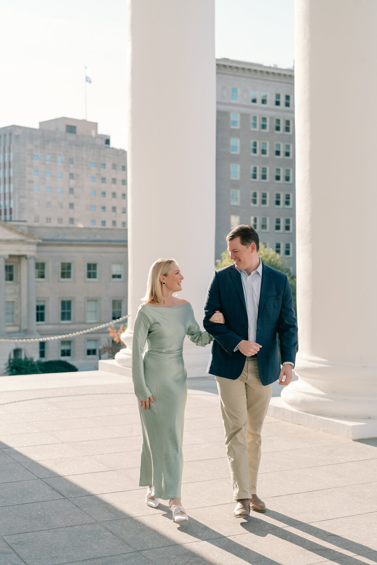 Virginia State Capitol Building Engagement Photos Richmond VA | newly engaged couple in green dress and blue suit walk arm in arm amidst the white columns of the VA Capitol Building