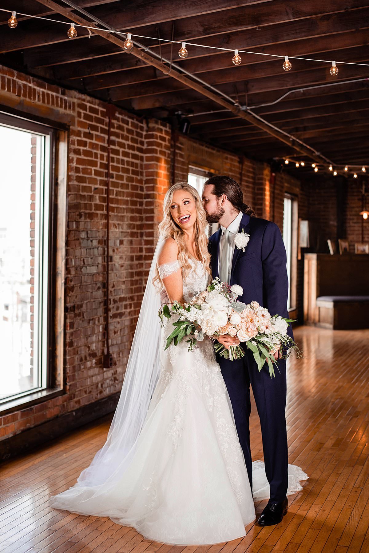Groom whispering into his fiances ear while she smiles and laughs, they are in indoor urban venue setting