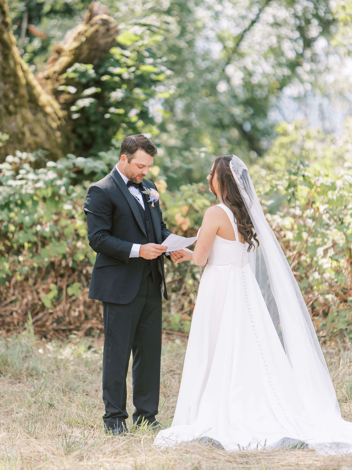 A groom holds the hand of his bride while he reads vows to her from a piece of paper at their outdoor luxury wedding.