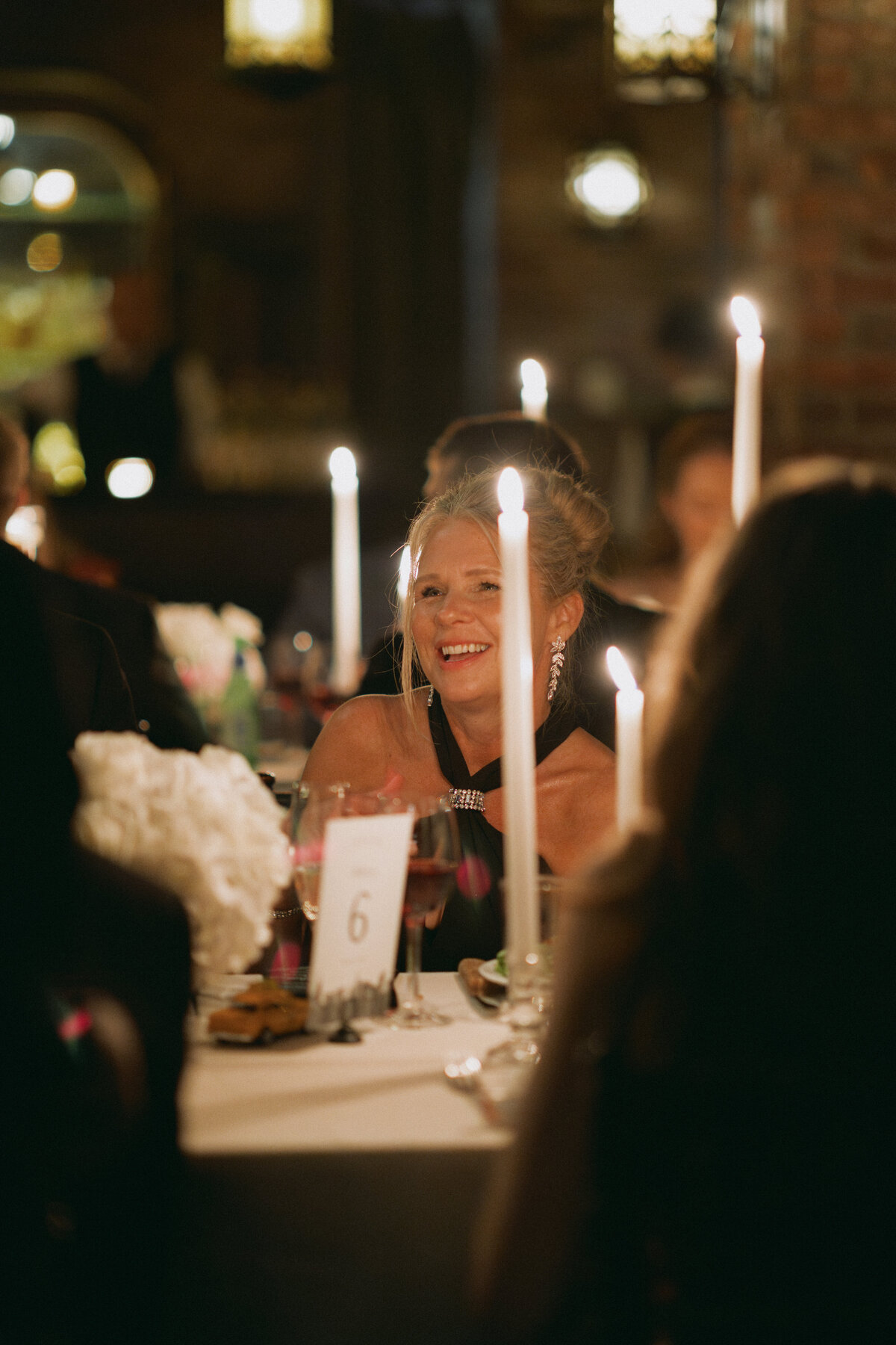 Wedding guest smiling during a candlelit reception dinner, surrounded by warm lighting, floral centerpieces, and glowing taper candles in an intimate and joyful celebration setting