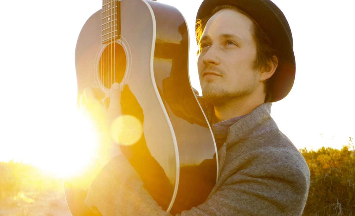 Country musician portrait Del Barber holding guitar high wearing tan coat black fedora while sun setting behind