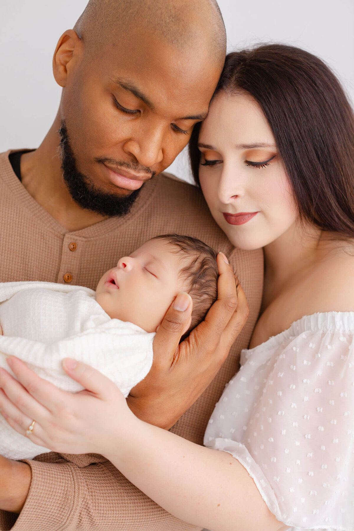 Dark-skinned dad and light-skinned mom hold newborn baby in their arms together while they are looking down at their new baby during newborn portraits.