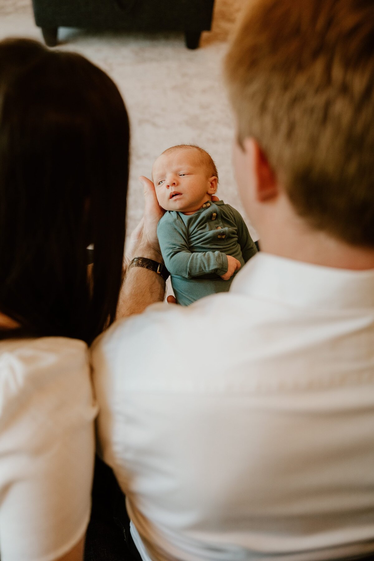 A newborn baby dressed in a green onesie is gently held and admired by his parents, with the focus on his tiny face as he gazes back, framed by the out-of-focus profiles of his mother and father.