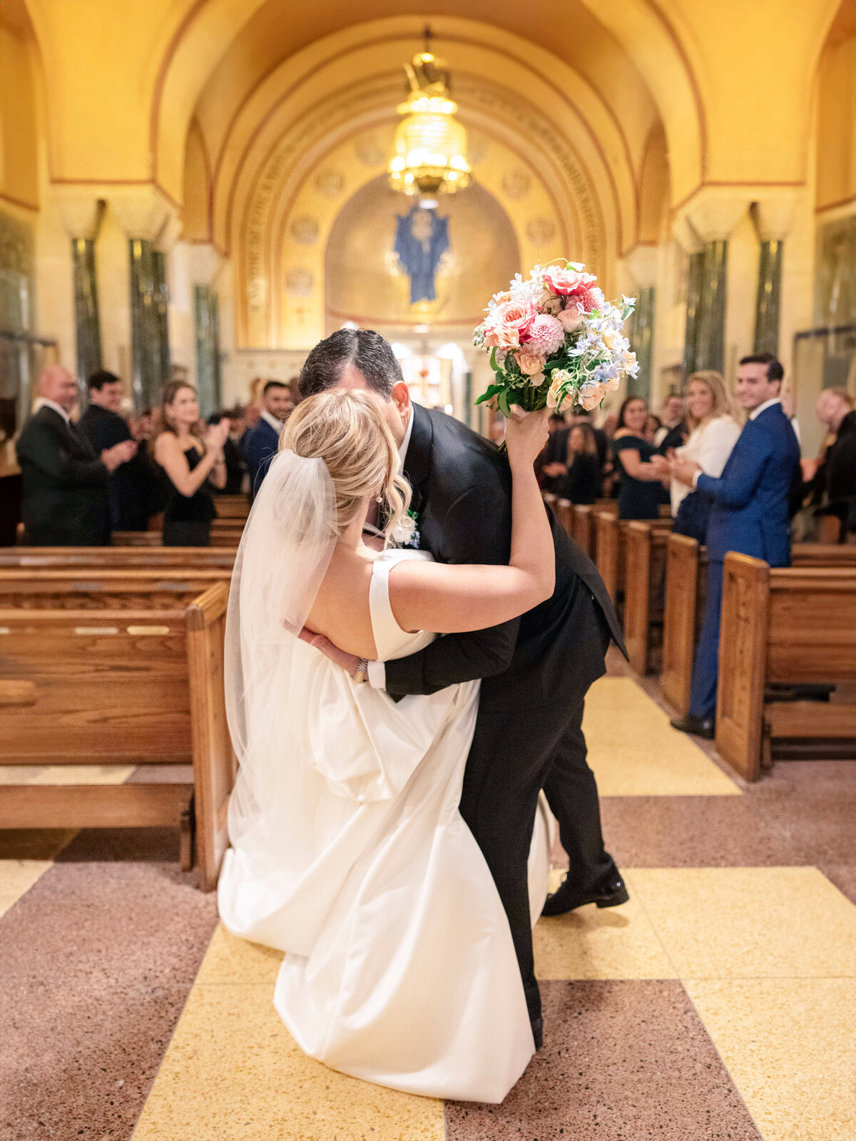 Bride and groom kiss in a church aisle as guests applaud. The bride holds a bouquet of flowers and wears a flowing white gown and veil. Guests are seated in wooden pews, and the church features an ornate arched ceiling.