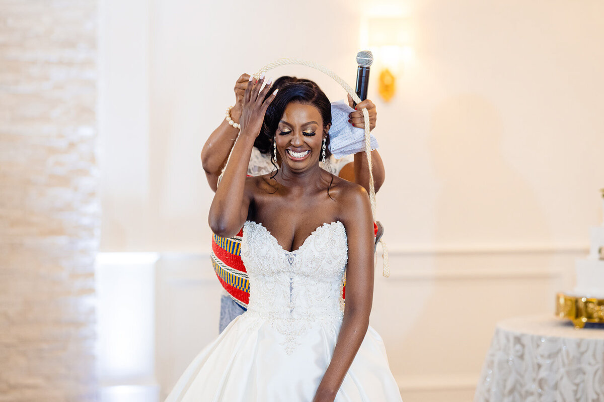 Bride in a wedding dress and tiara joyfully dancing with a Kente cloth being placed on her shoulders