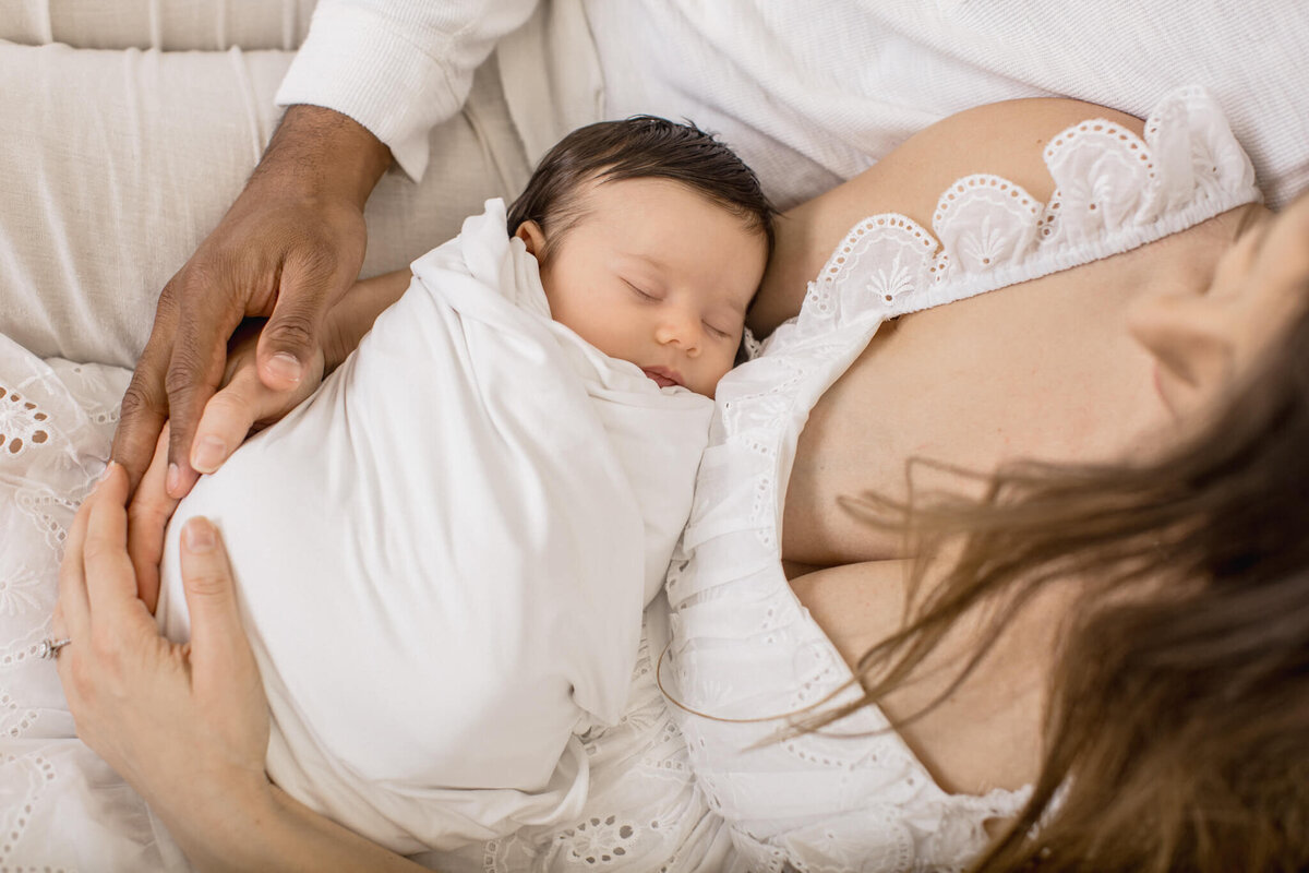 A newborn baby sleeps peacefully wrapped in a white blanket. The baby is cradled in the arms of an adult wearing a white lace dress. Another adults hand rests gently on the baby, also wearing a white shirt, creating a serene and tender scene.