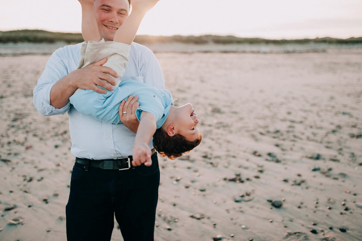 A dad playfully flips his son upside down on the beach at sunset