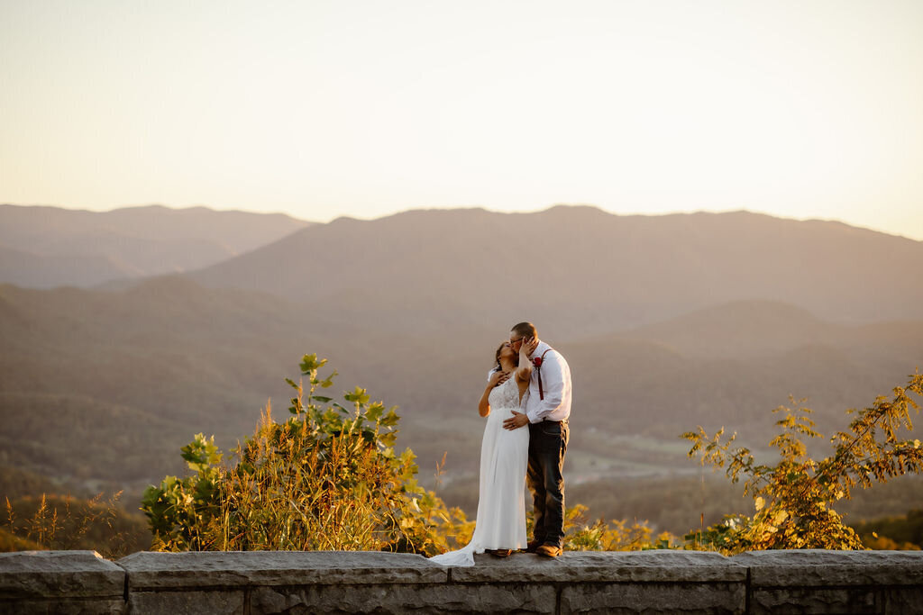 Foothills Parkway wedding photos with couples eloping to Gatlinburg standing on a stone wall with the mountains in stand distance as they kiss 