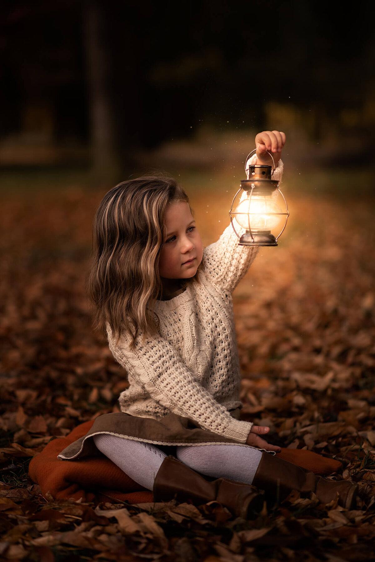 Fall in Kamloops features a little girl sitting on the ground in the leaves holding up a glowing lantern while looking into it.
