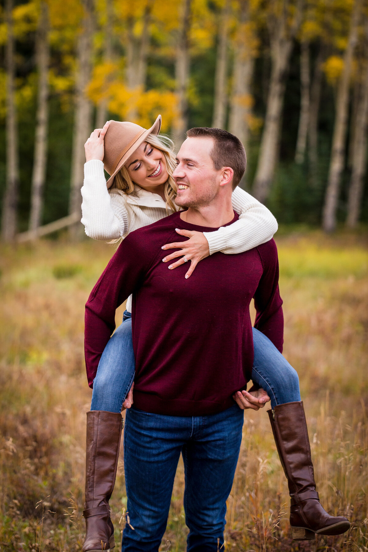 A man gives his fiancée a piggyback ride as she smiles and holds onto her hat in a fall setting.