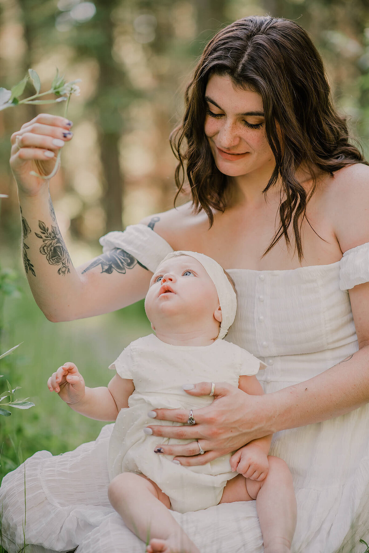A mother and daughter sit in the tall grass together. Baby girl is on moms lap looking up at the piece of grass mom is holding in her hands. Mom smiles down at her. Taken by Brittany Danielle Photography