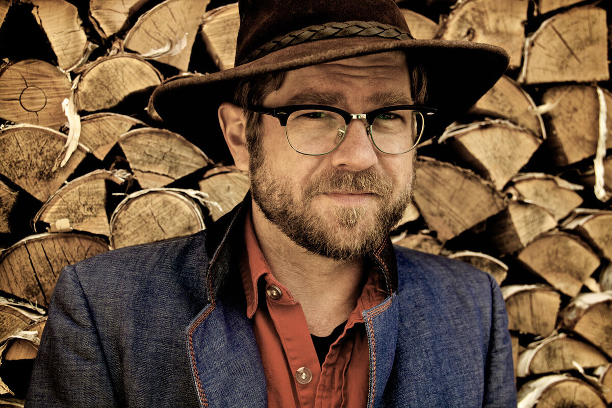 Country Music Portrait Old Man Luedecke standing in front of wood pile wearing wide brim hat and glasses