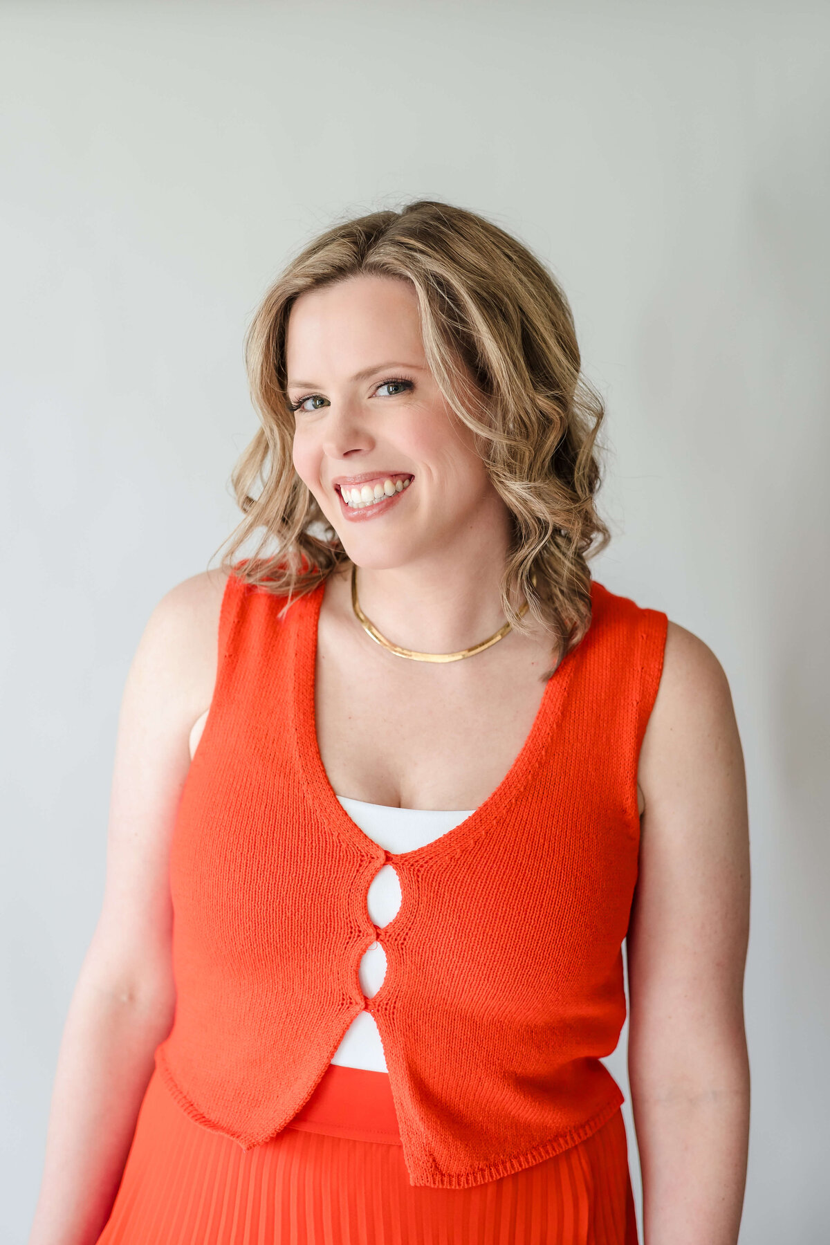 Brittany, a naturopathic physician, smiling in orange knit top and gold necklace at Base Coat Studios.