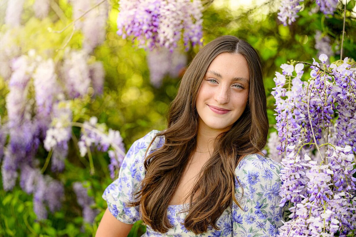 Charlotte Catholic graduate sits in a blue and purple floral dress surrounded by wisteria for her senior portraits