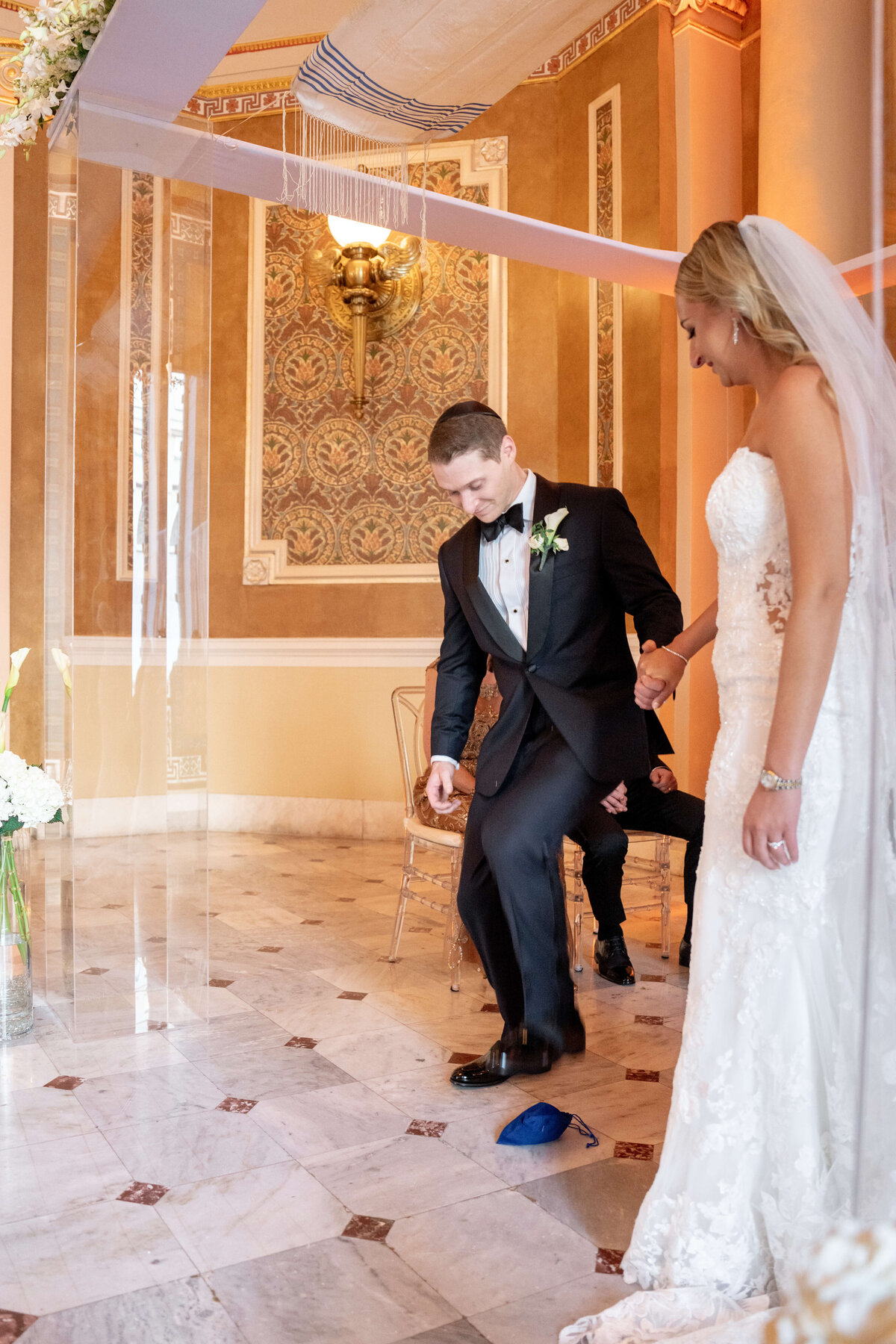 A Jewish groom in a tuxedo stomps on a glass, likely a glass, under a canopy during a wedding ceremony. The bride in a white dress stands beside him, holding his hand. The setting is an ornate room with brown and gold decor.