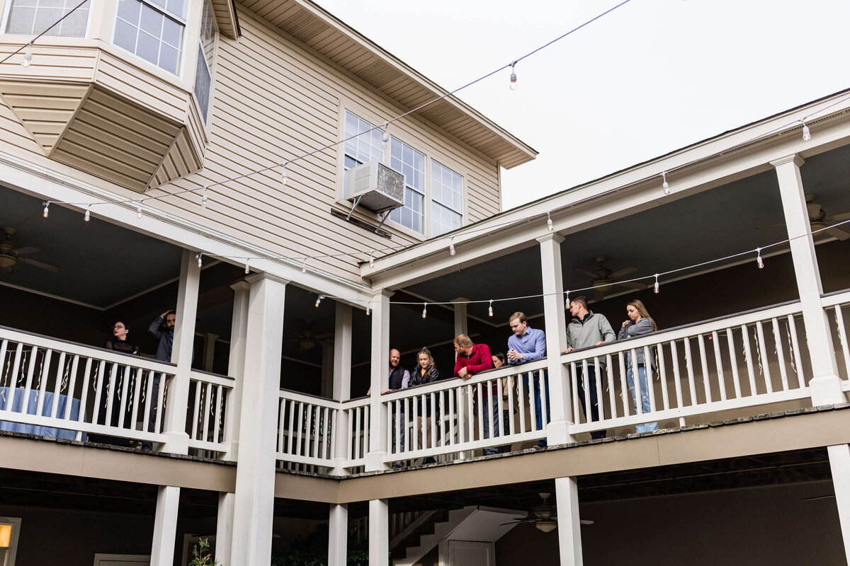 A group of people stand and lean on the railing of an outdoor balcony at a two-story building, talking and looking down, with string lights hanging above them.