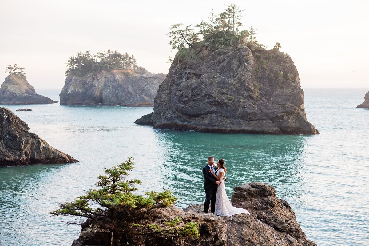 A bride and groom stand on a cliff edge overlooking secret beach. 